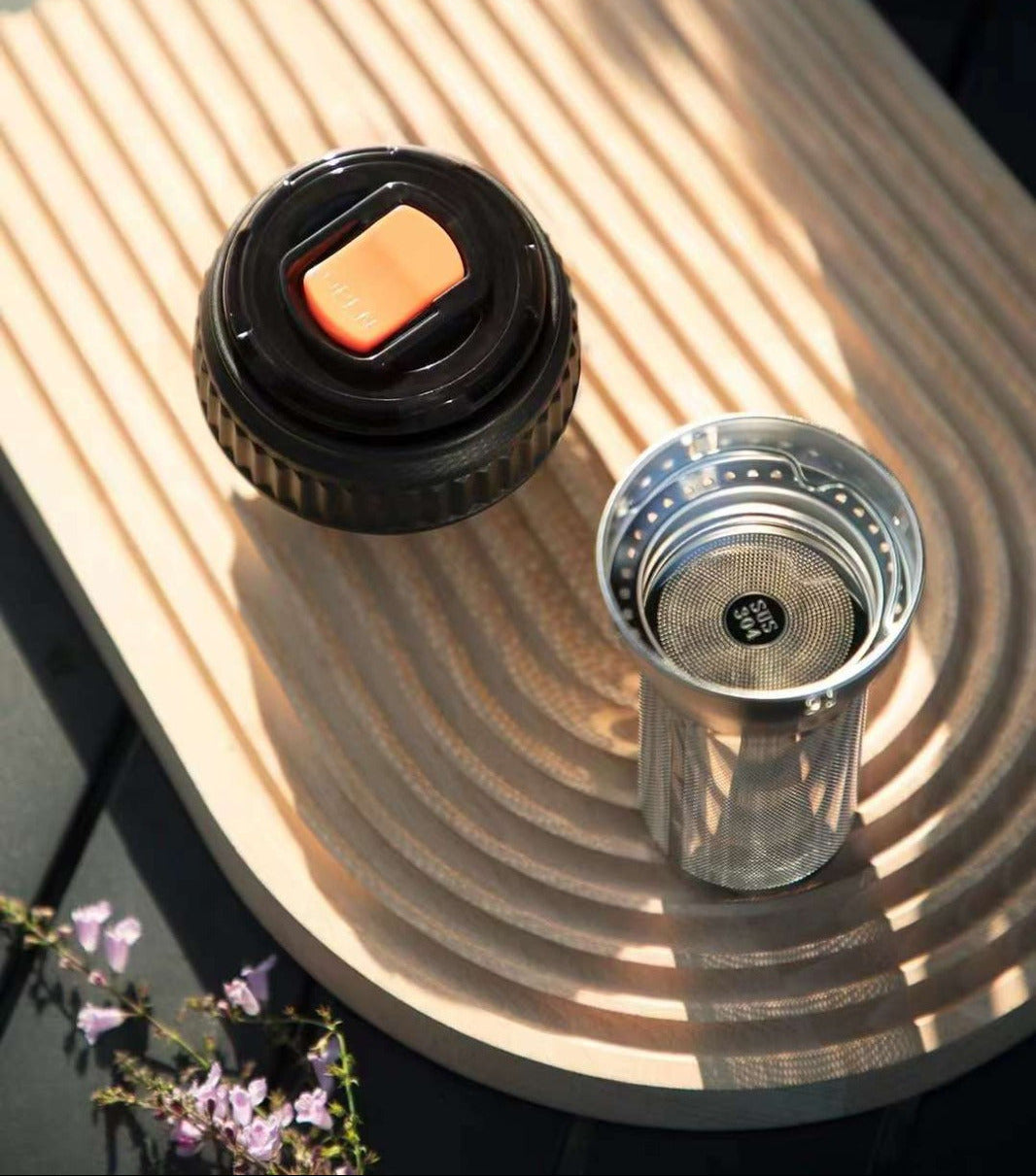 A close-up product shot of a tea infuser and lid set on a curved wooden tray. The set includes a black lid with a clear plastic plug and an orange accent, and a stainless steel tea infuser cylinder.